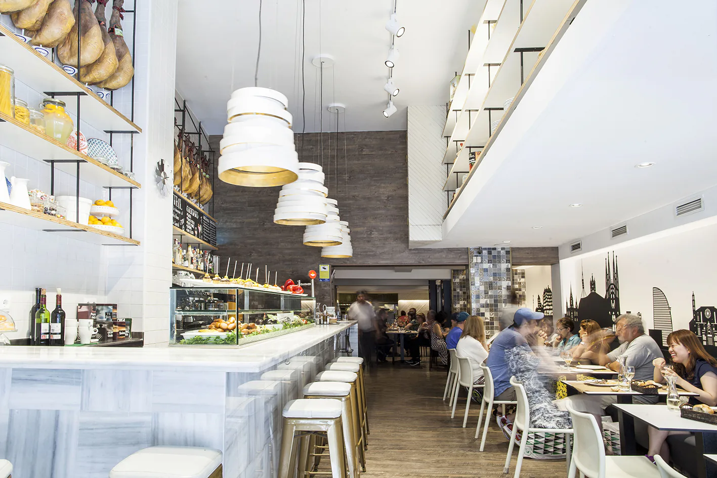 La barra de mármol con los jamones colgados encima, y al fondo el comedor con clientes comiendo.