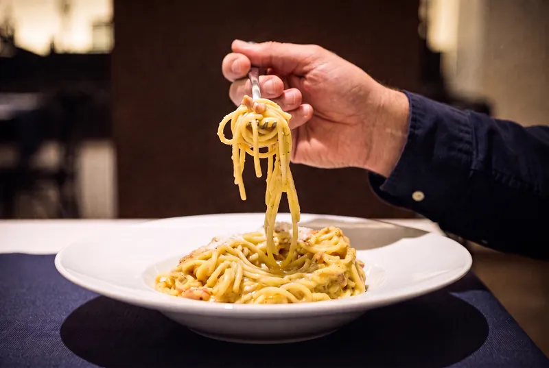 Una forquilla aixeca una forquillada d’espaguetis a la carbonara, enrotllats, per sobre del plat fondo.