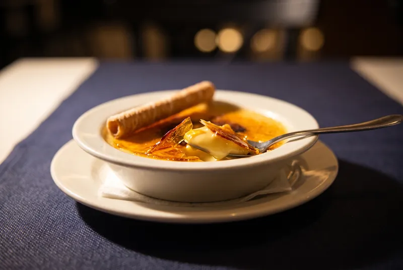 Una cullera trenca el caramel d’una crema catalana, amb la seva neula al damunt, en un bol blanc sobre les estovalles blaves.