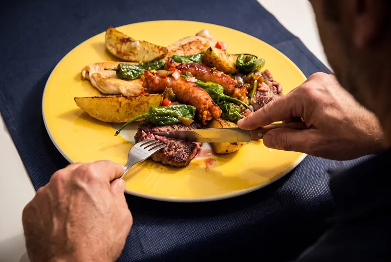 Algú talla la vedella d’una graellada de carn: botifarra, pit de pollastre, pebrots verds i patates en un plat groc.