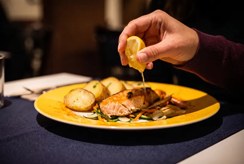 Una mà esprem un grill de llimona sobre un salmó a la planxa, amb patates i verduretes en un plat groc.