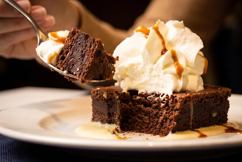 Una cullera s’endú el primer tros d’un pastís de xocolata amb nata muntada, caramel i crema anglesa.