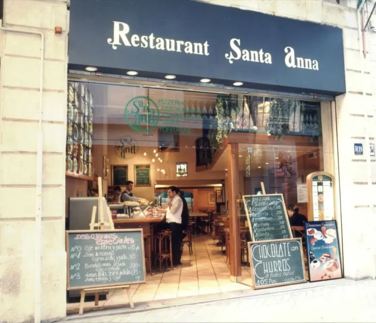 La fachada con el rótulo azul Restaurant Santa Anna y dos pizarras en la acera con los desayunos y los churros con chocolate en pesetas.