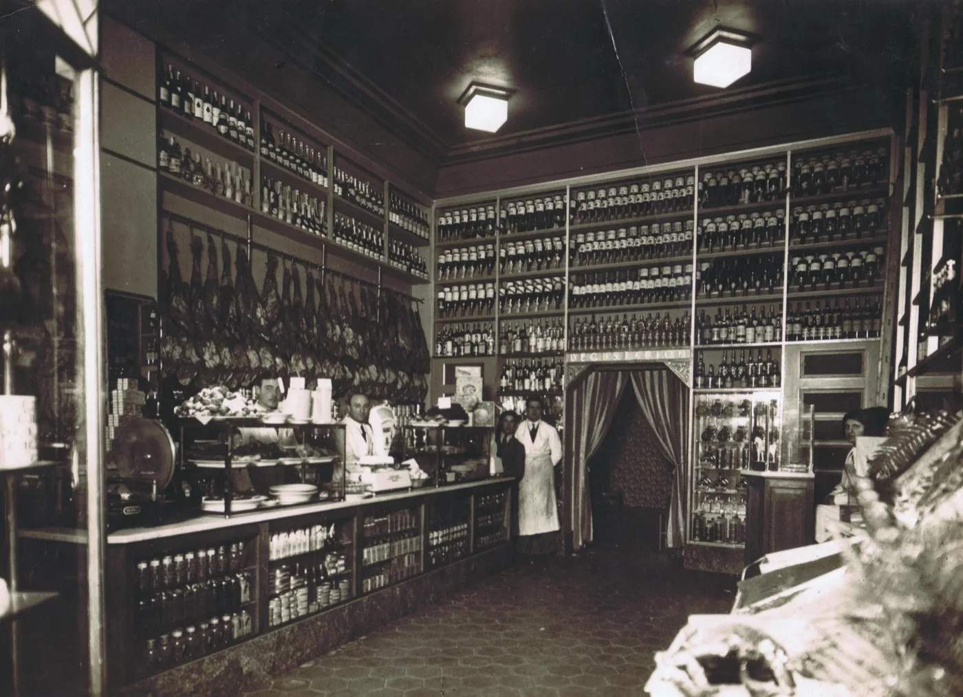 Interior antiguo del número 8: jamones colgados, estanterías de conservas y botellas, y dos empleados detrás de un mostrador de mármol.