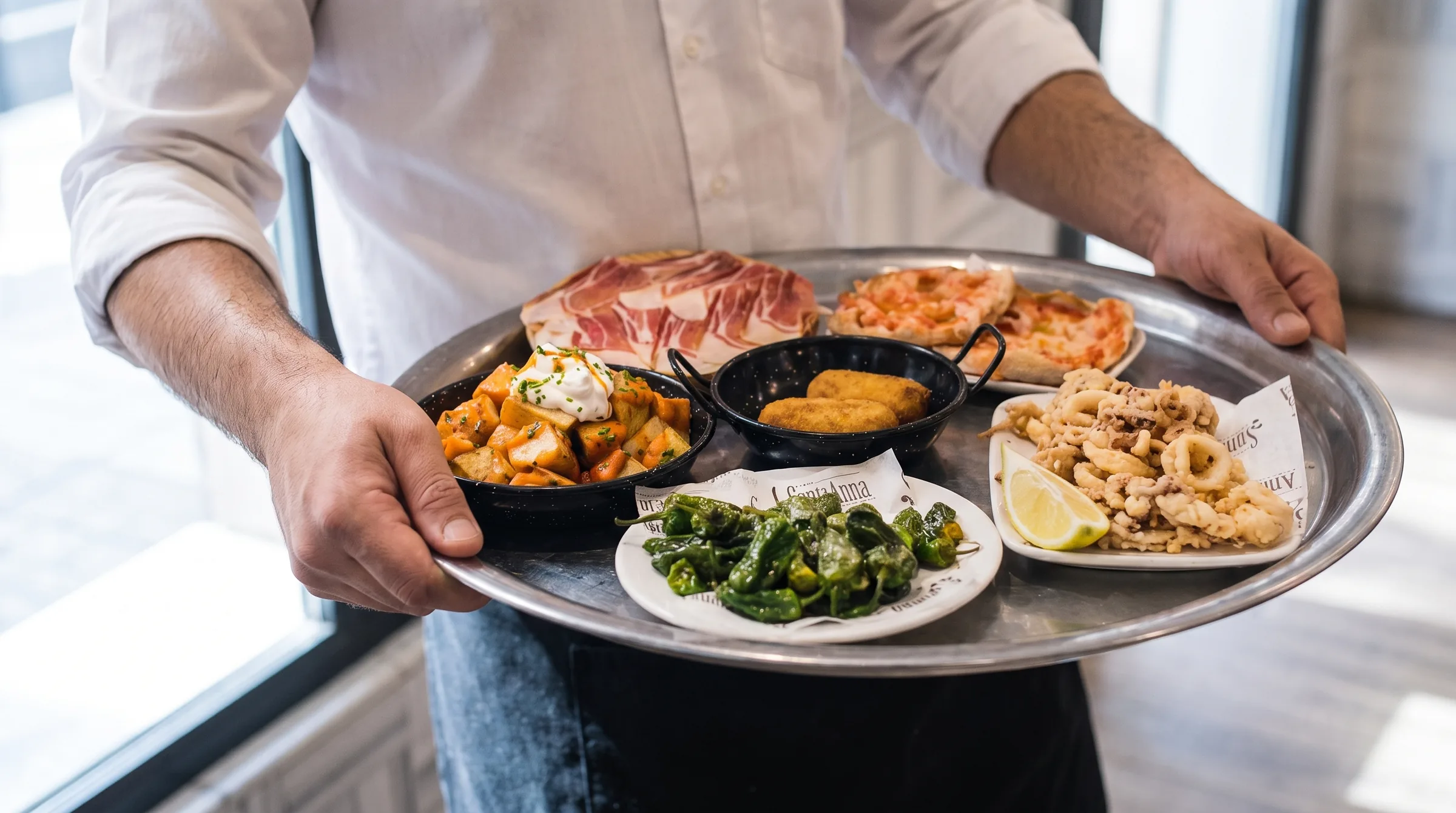Un cambrer porta una safata amb sis tapes: patates braves, croquetes, pebrots del Padró, pa amb tomàquet, calamars i pernil.