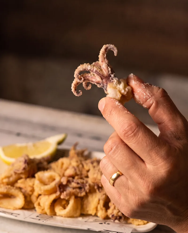 Una mà sosté un tentacle de calamar fregit, amb el plat i el grill de llimona al darrere.