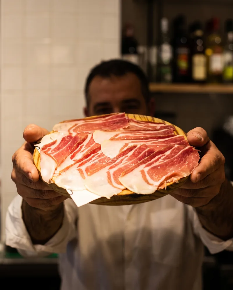 Un cambrer acosta la post de pernil ibèric, amb les ampolles de la barra al fons.