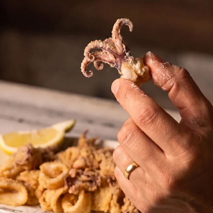 A hand holding up one fried squid tentacle, the plate and the lemon wedge behind it.