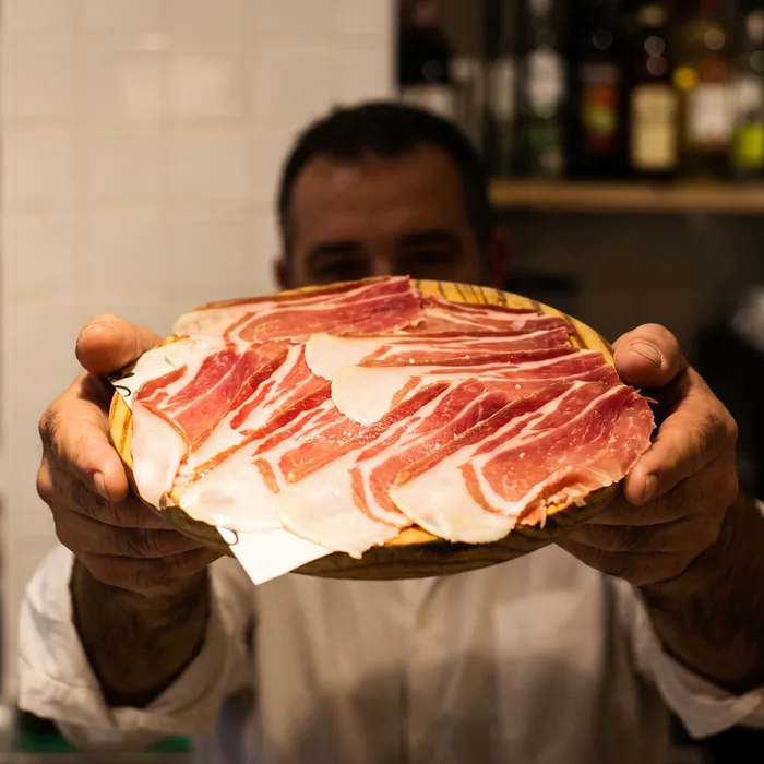 A waiter holding out the board of Iberian ham, with the bar’s bottles behind.