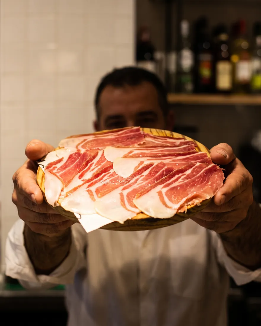Un camarero acerca la tabla de jamón ibérico, con las botellas de la barra al fondo.